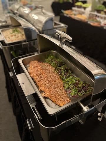 Buffet tray of seasoned roasted fish garnished with herbs under a warming lid
