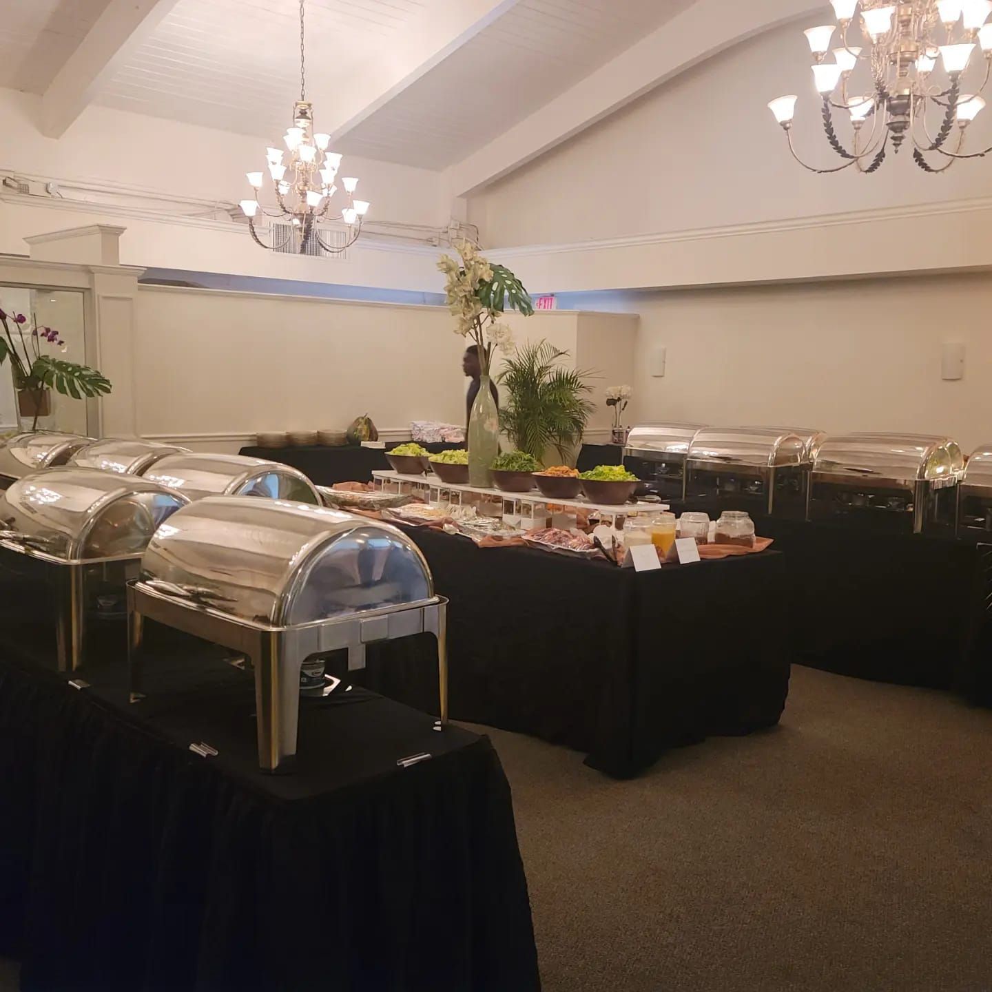 Buffet tables with covered chafing dishes and trays in a banquet hall, lit by chandeliers