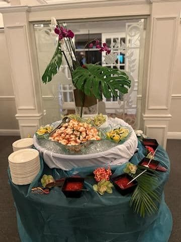 Tropical buffet table with seafood platter, fruit, and flowers under large green leaves in a banquet hall