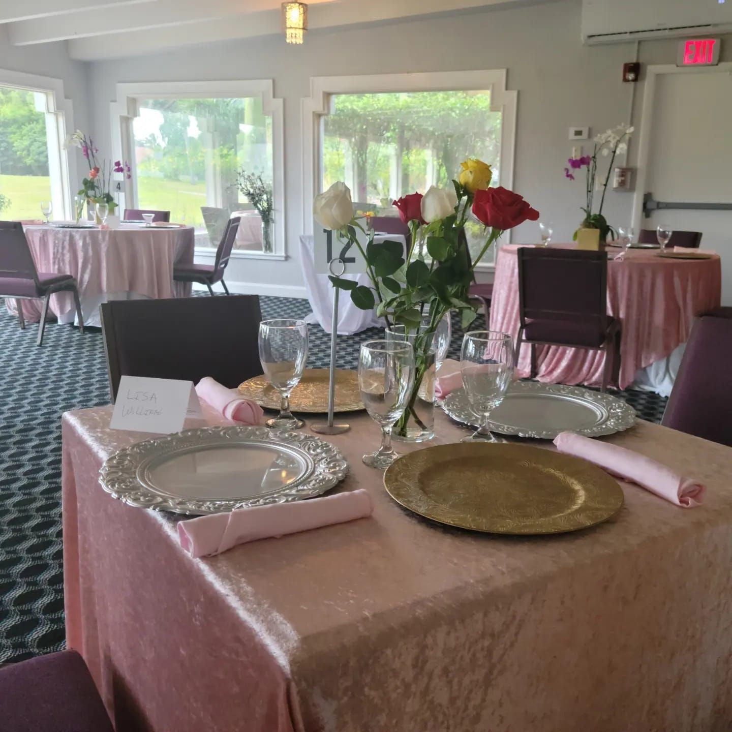 Set banquet table with pink tablecloth, silver plates, folded napkins, glassware, and floral centerpiece in a bright room