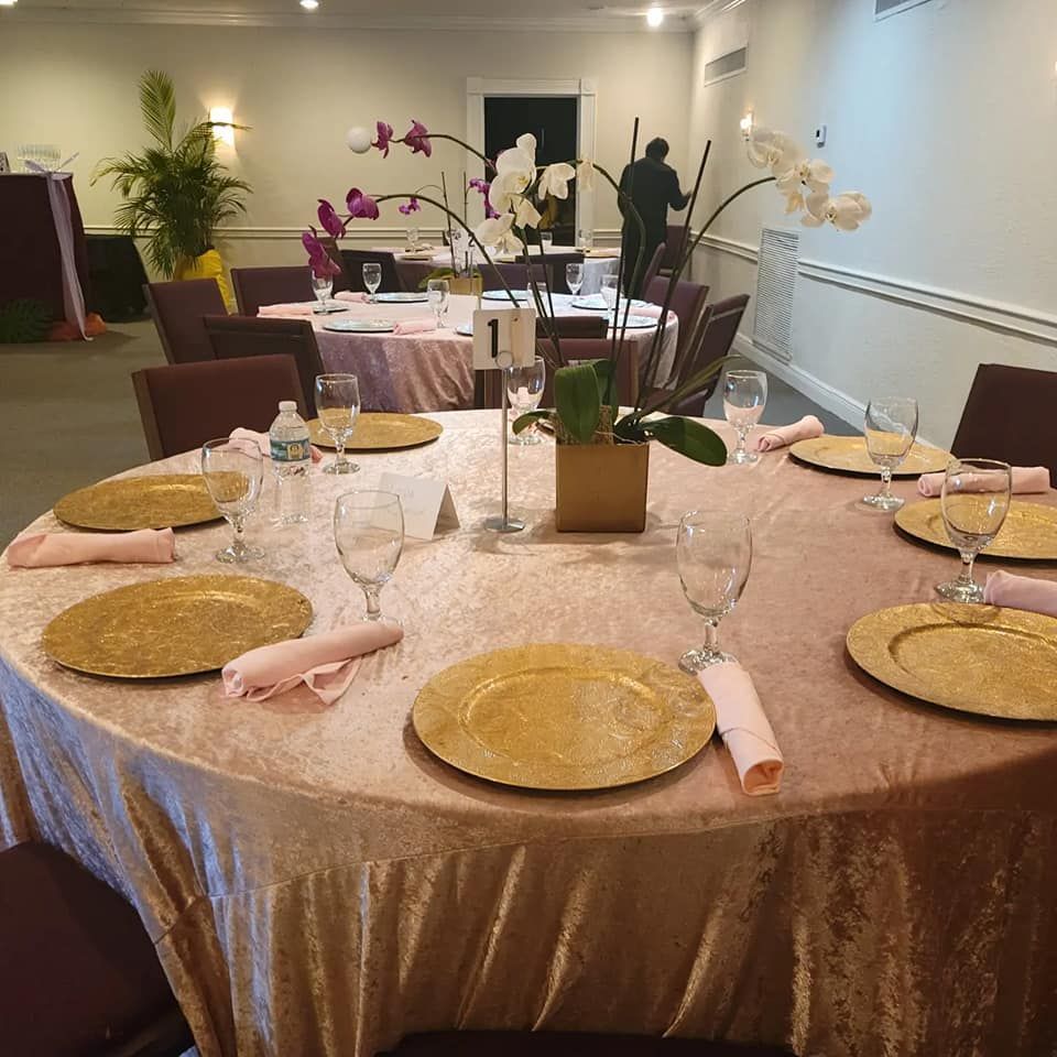 Round banquet table set with pink napkins, gold plates, wine glasses, and floral centerpieces in a hall