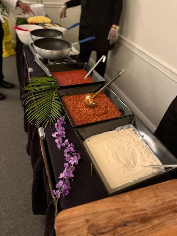 Buffet table with pans of sauces and a tray of rice beside decorative flowers and palm leaves