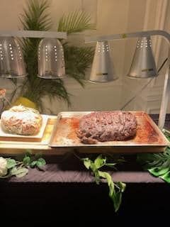 Buffet table with covered dishes, a loaf of bread, and a tray of roasted meat under warming lights