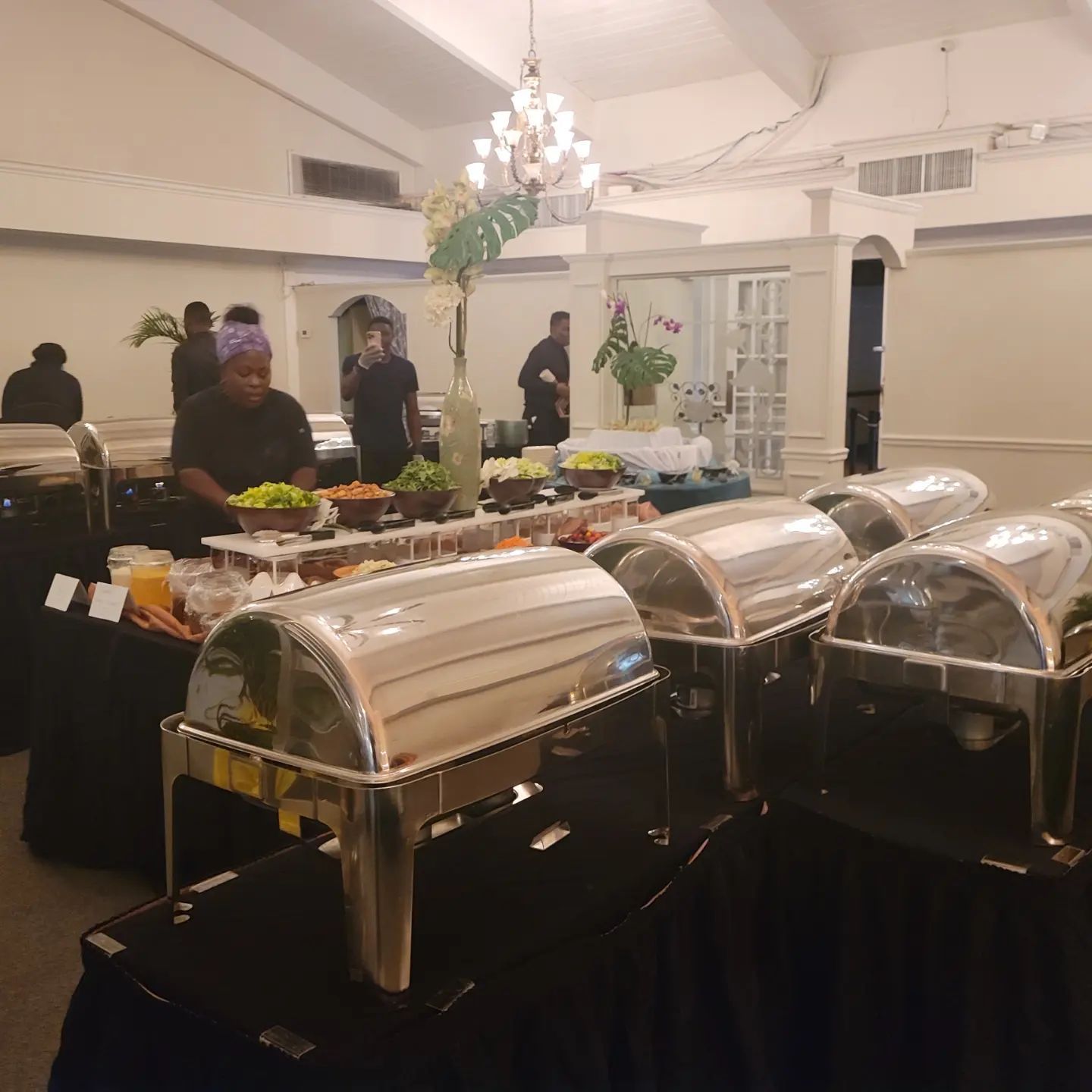 Buffet setup in a banquet hall with covered chafing dishes and guests near a decorated table