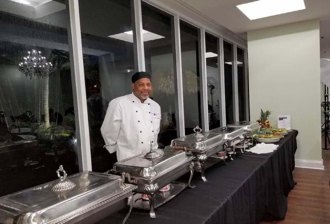 Chef in a white jacket holding two plated dishes in a commercial kitchen.