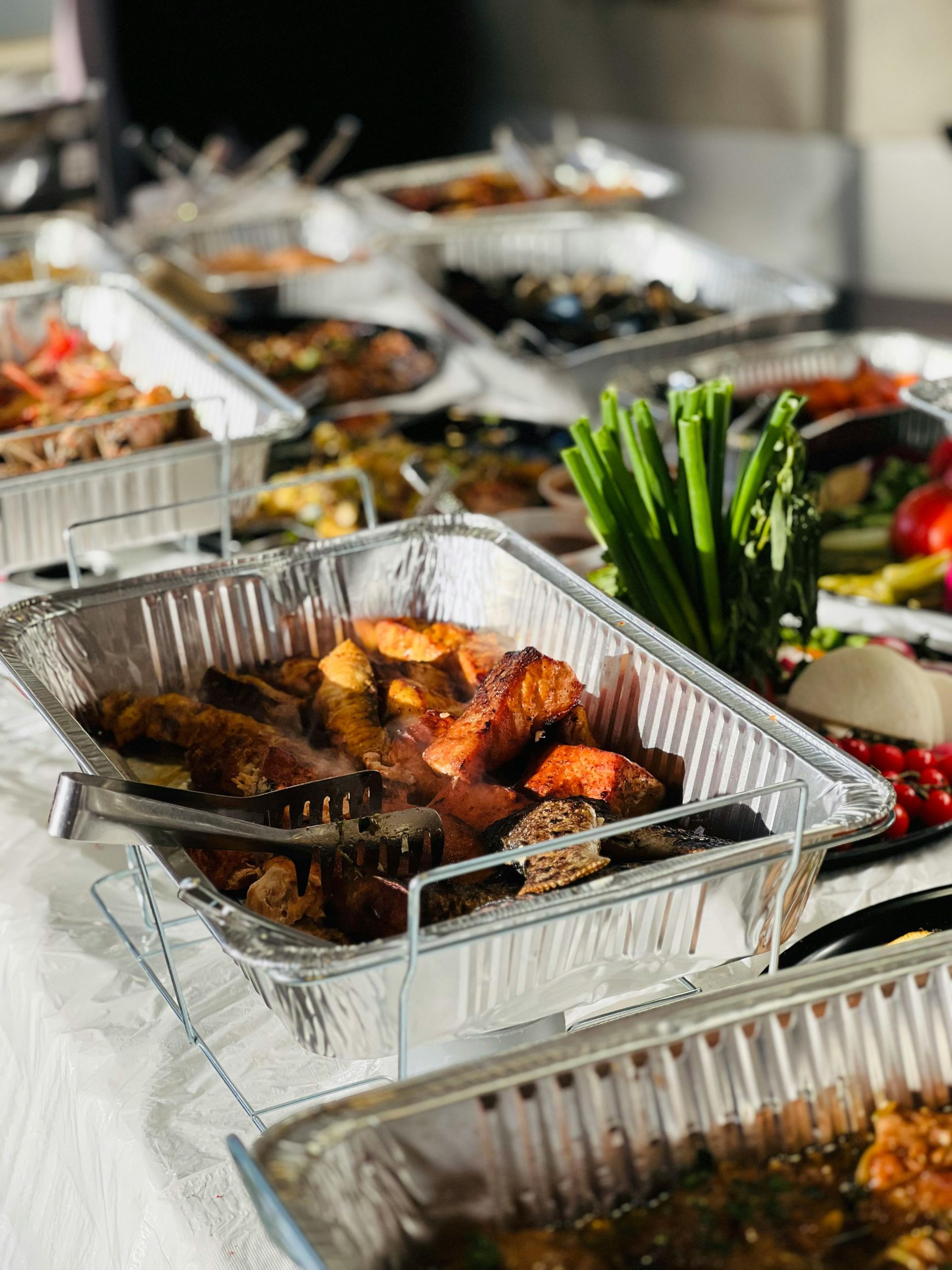 Tray of grilled meat and vegetables at a buffet, with multiple foil pans of food in the background
