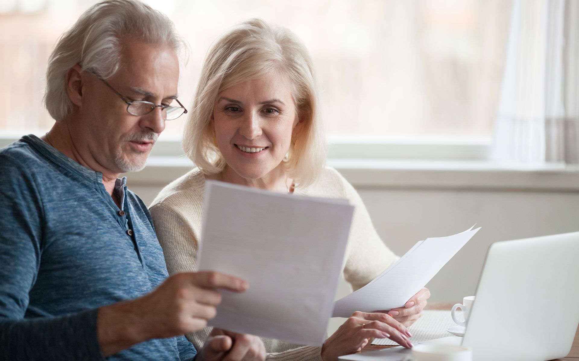 An elderly couple is sitting at a table looking at papers and a laptop.