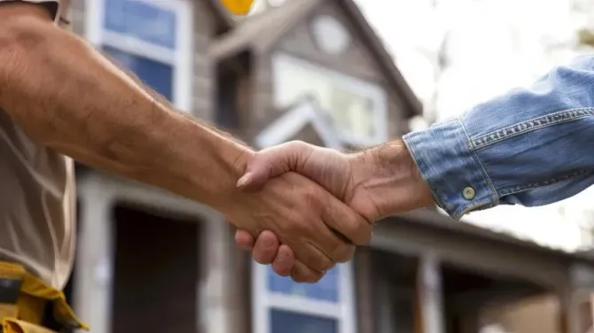 Two people shaking hands in front of a house under construction.