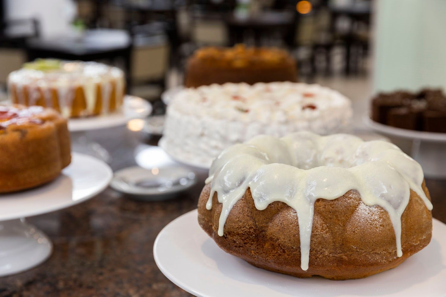 Um bolo bundt com cobertura branca está sobre uma mesa com outros bolos.