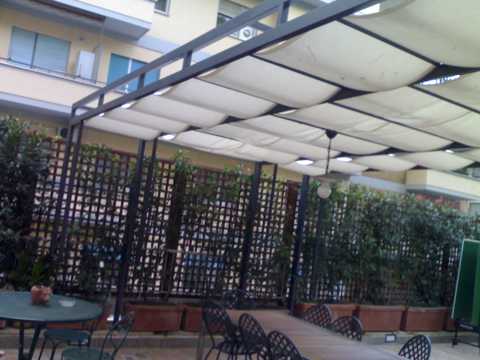 Patio esterno con tavoli e sedie in metallo nero sotto una tettoia bianca accanto a un edificio