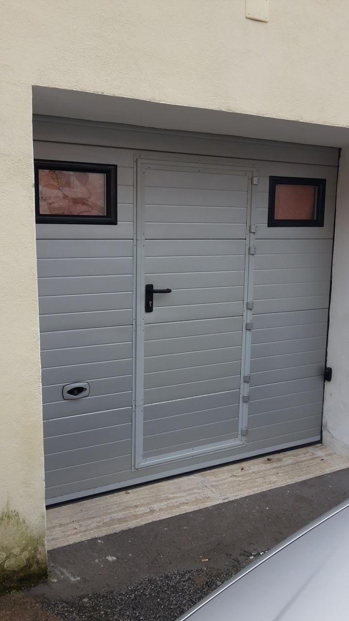 Porta da garage grigia con due piccole finestre in una parete beige, vista dalla strada.