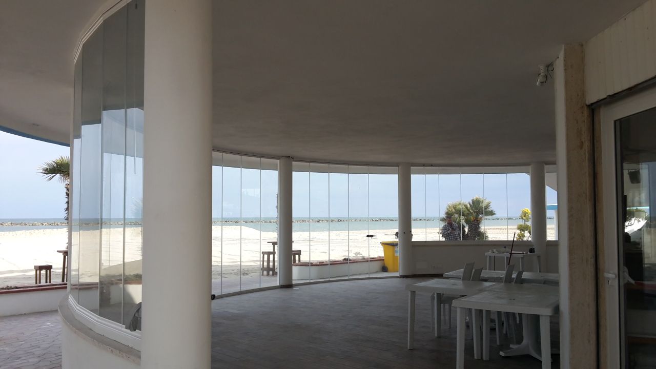 Patio coperto fronte mare con colonne bianche, ringhiera in vetro e tavoli all'aperto con vista sull'oceano.