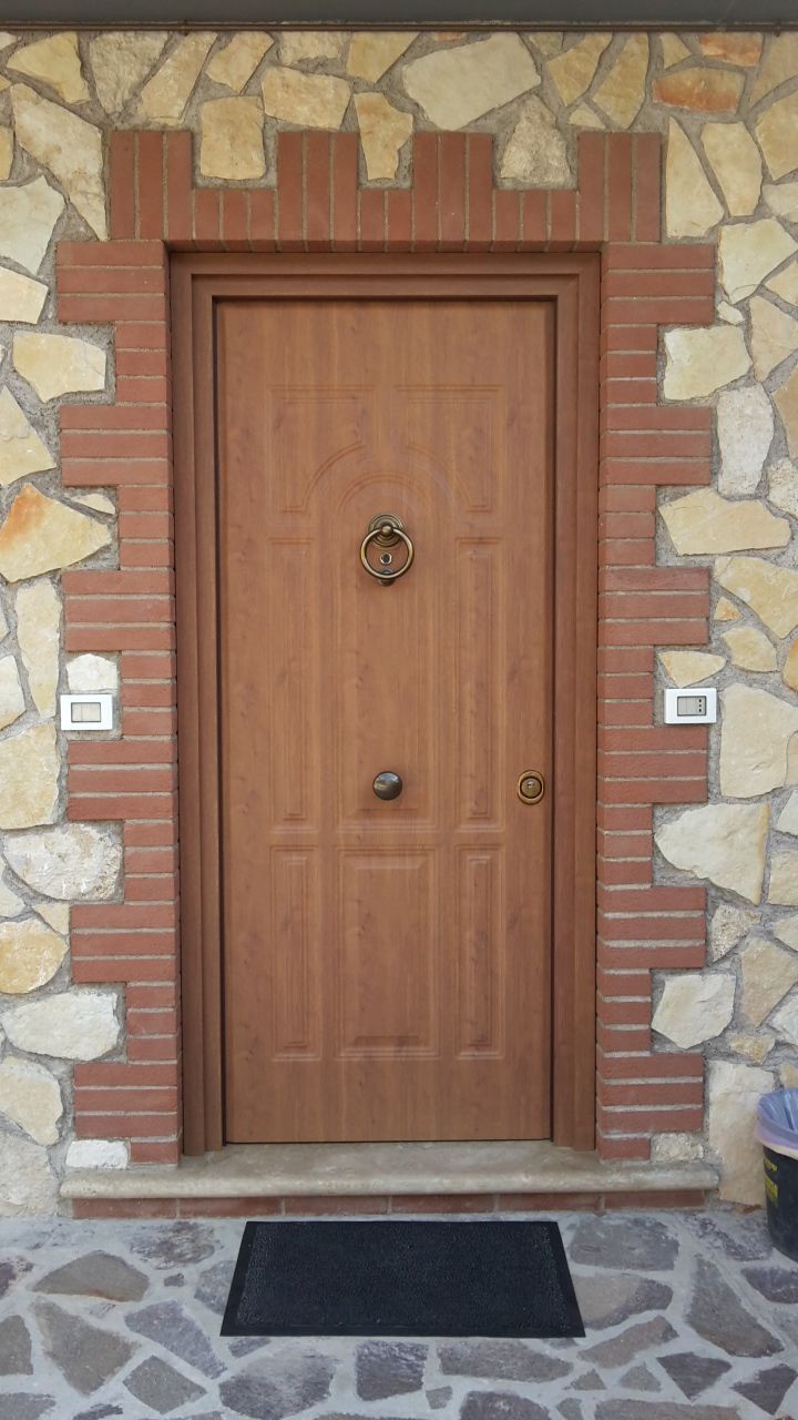 Porta d'ingresso in legno incastonata in una facciata in pietra con zerbino nero e targhette laterali