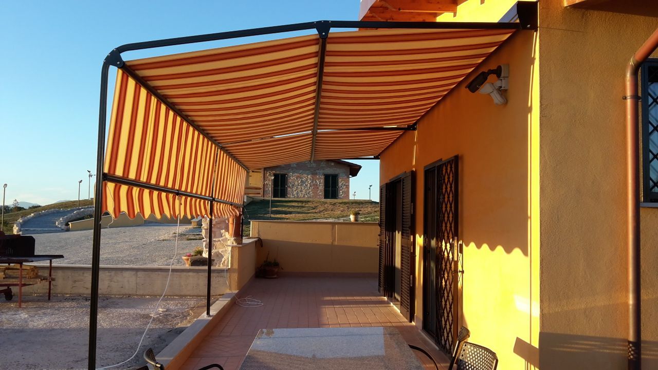 Patio di una casa gialla con tenda da sole a righe che proietta ombre su un vialetto piastrellato e una ringhiera.