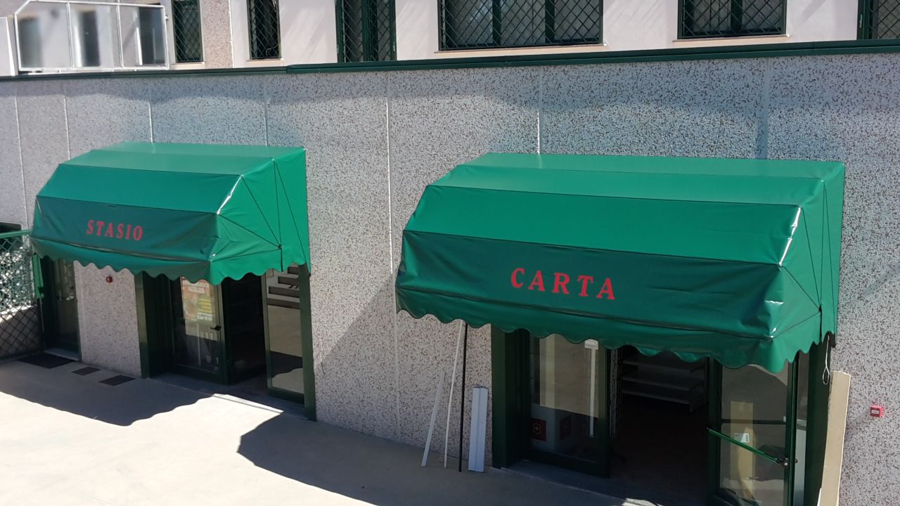 Due tende da sole verdi sopra gli ingressi dei negozi su un edificio grigio, una delle quali reca la scritta