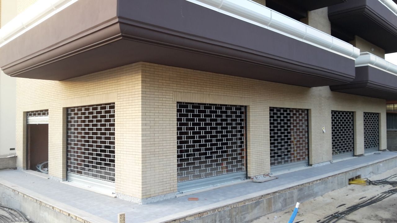 Angolo di un edificio in mattoni beige con griglie metalliche scure sopra le aperture del piano terra e un tetto sporgente.