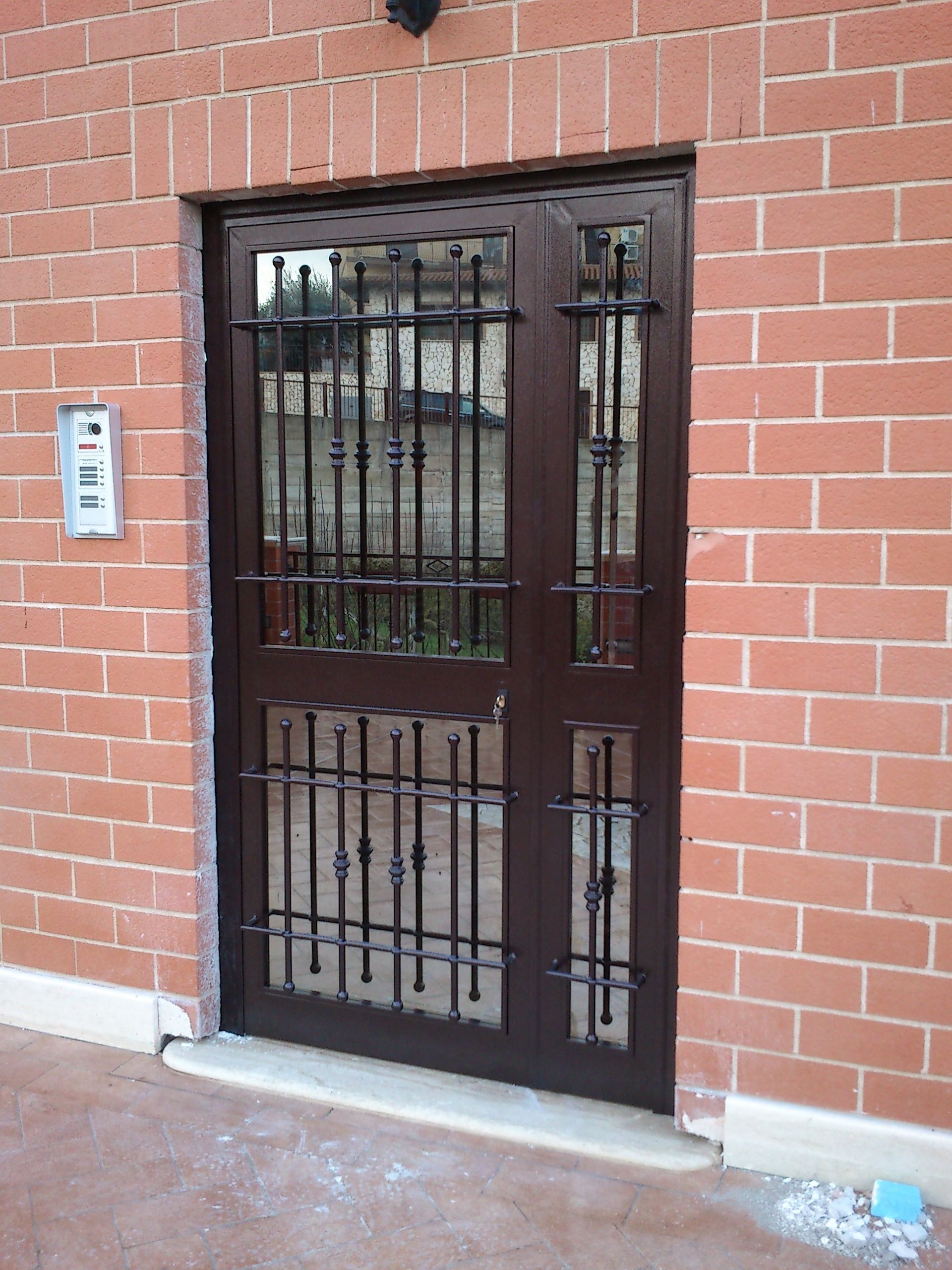 Porta d'ingresso in vetro incastonata in un muro di mattoni rossi con sbarre in metallo nero e pannello di ingresso con tastiera numerica.