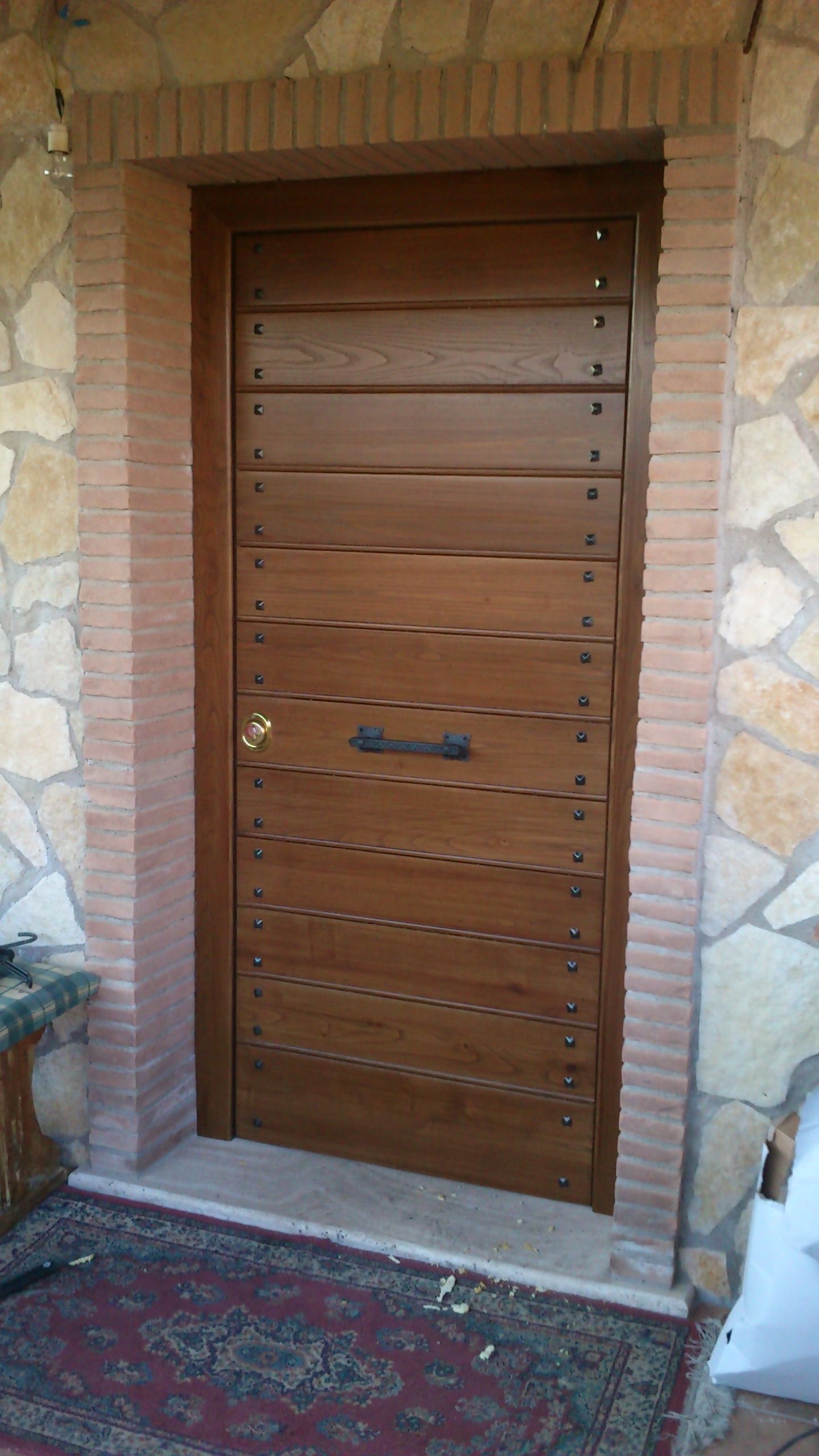 Porta d'ingresso in legno con doghe orizzontali, inserita in un portale in pietra e mattoni, con zerbino sottostante.