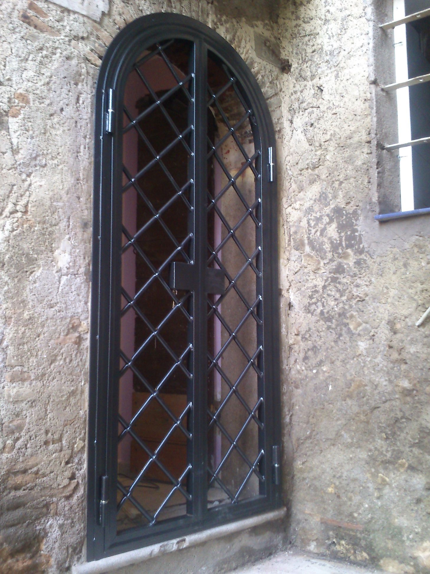Stretto cancello ad arco in metallo nero incastonato in un muro di pietra consumato dal tempo.