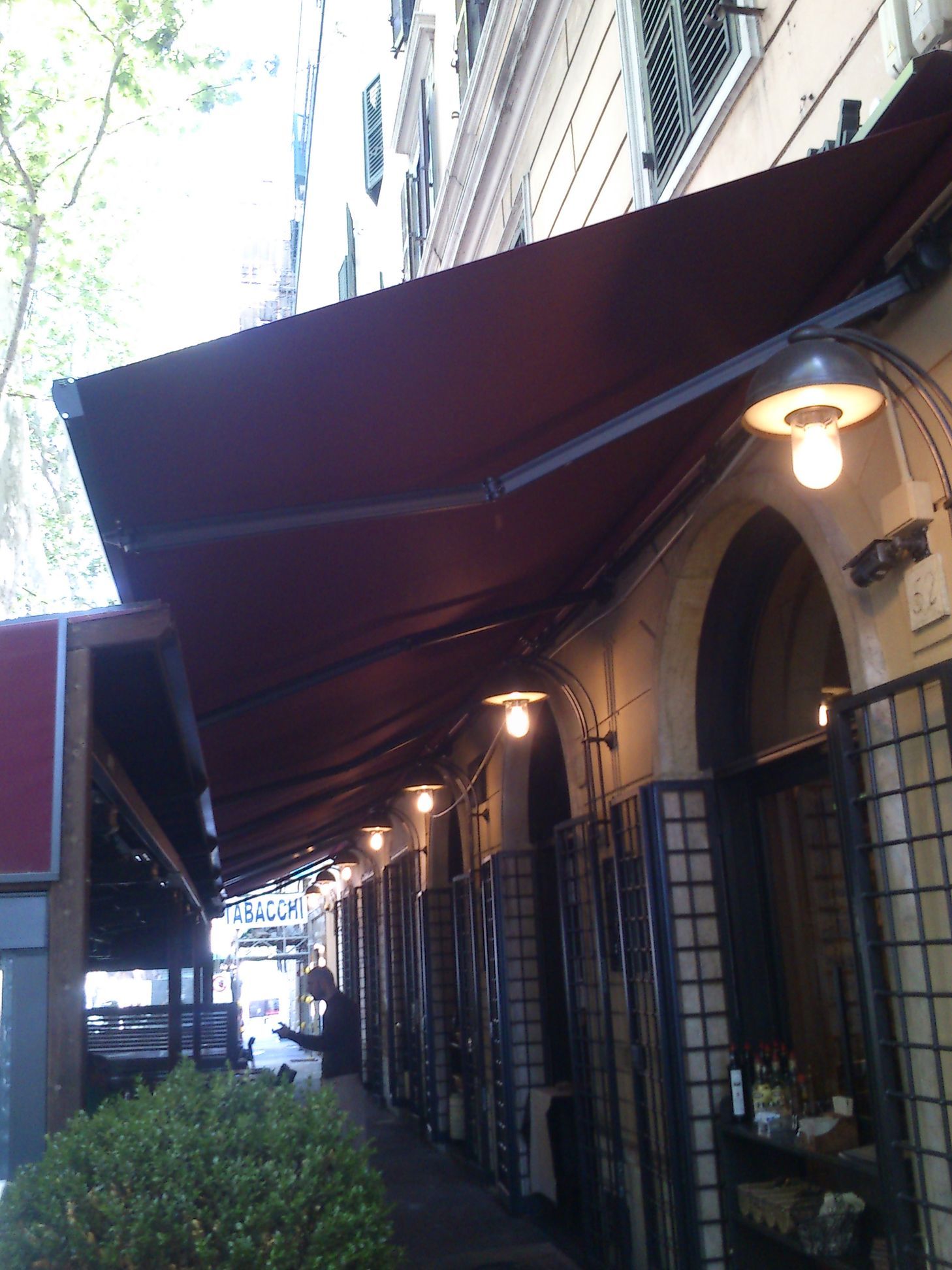 Tenda da sole color bordeaux all'esterno di uno stretto marciapiede antistante un negozio, con lampade accese e finestre ad arco.