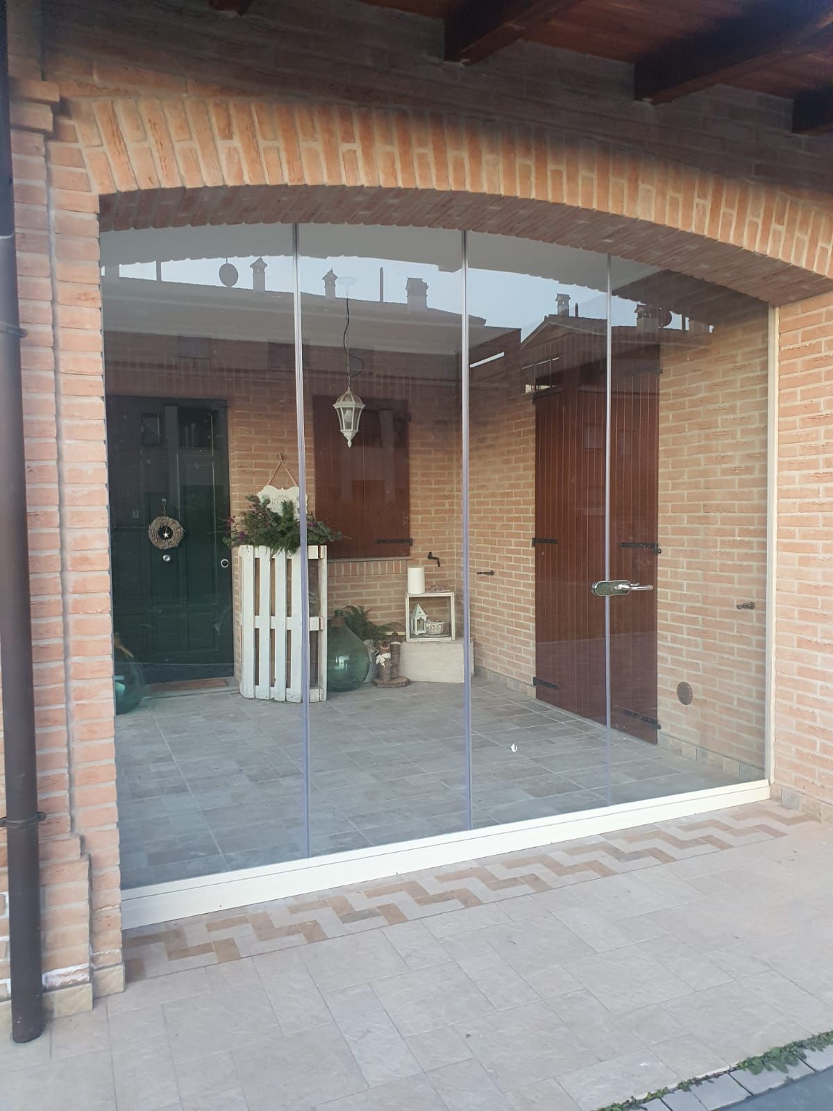 Ingresso di una casa in mattoni con porte anteriori in vetro, un portico coperto e piante in vaso su un pavimento piastrellato.