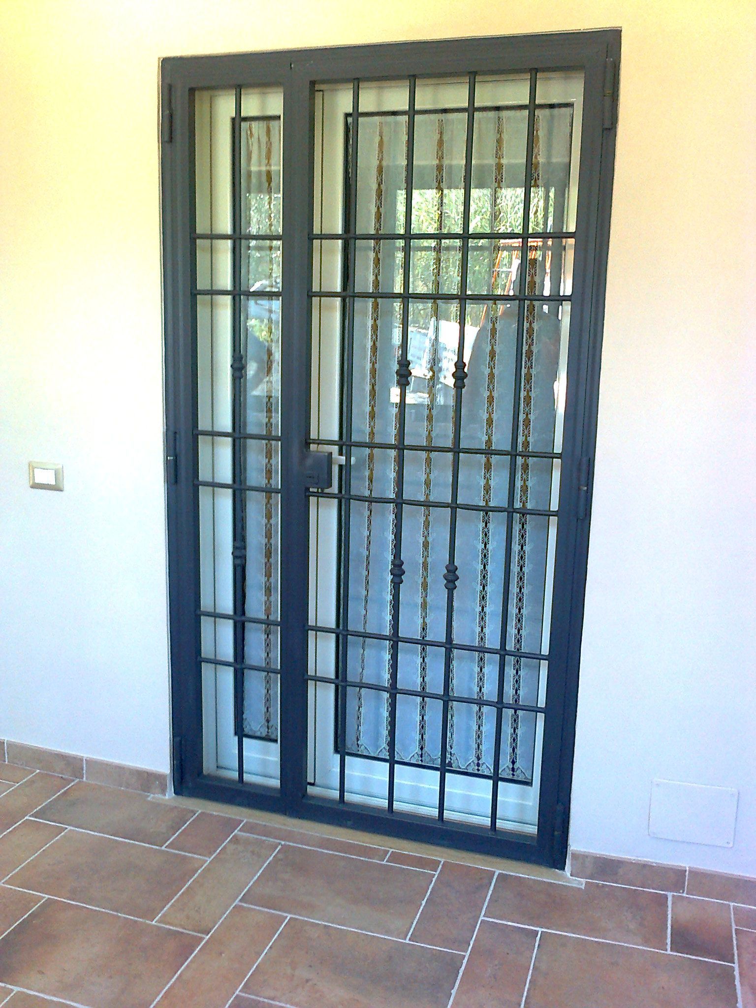 Porta blindata in metallo nero con pannelli in vetro e sbarre, installata su un muro piastrellato del patio.