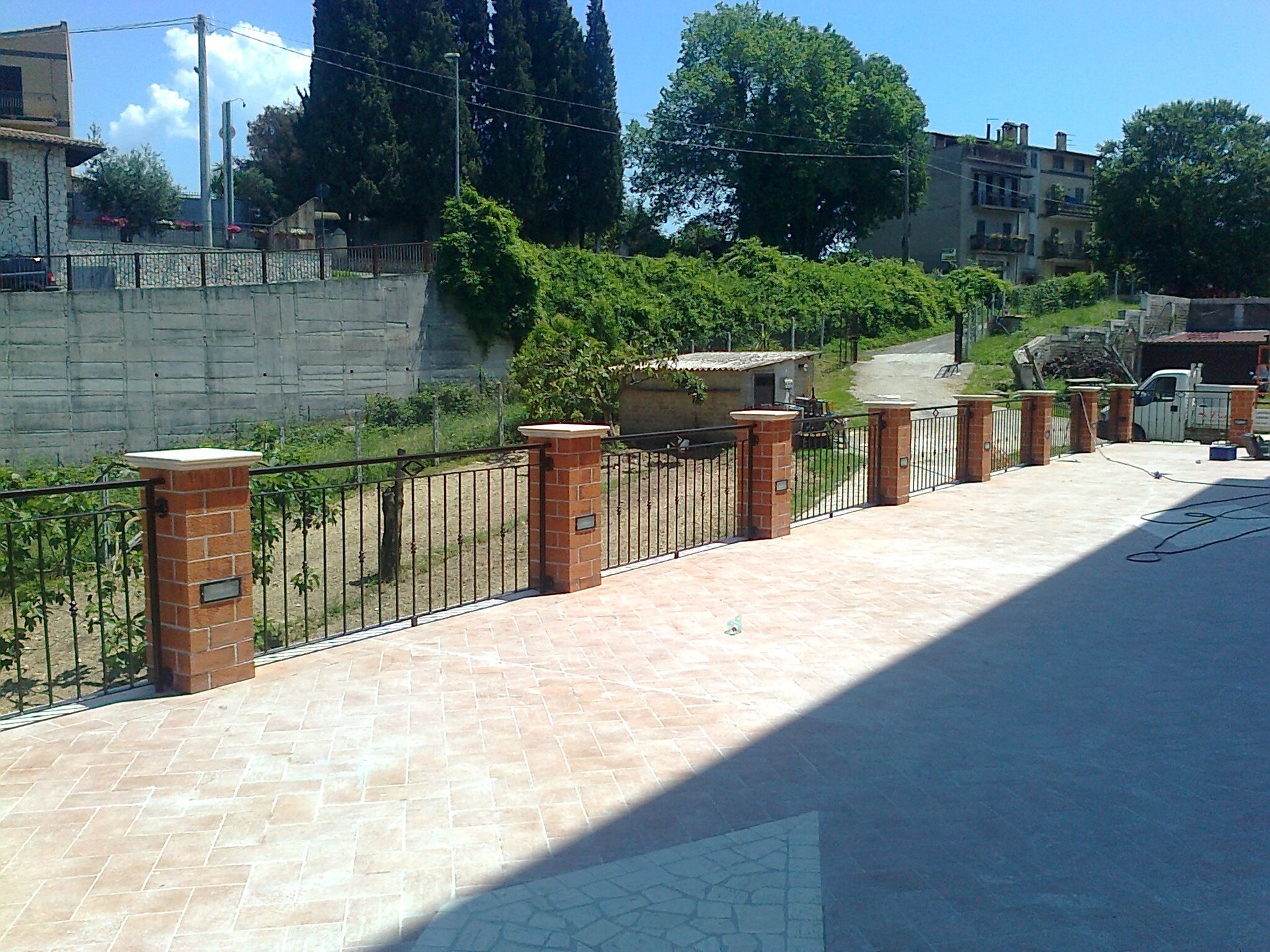 Passeggiata lastricata e soleggiata con pilastri in mattoni e siepe verde adiacente a una zona residenziale.
