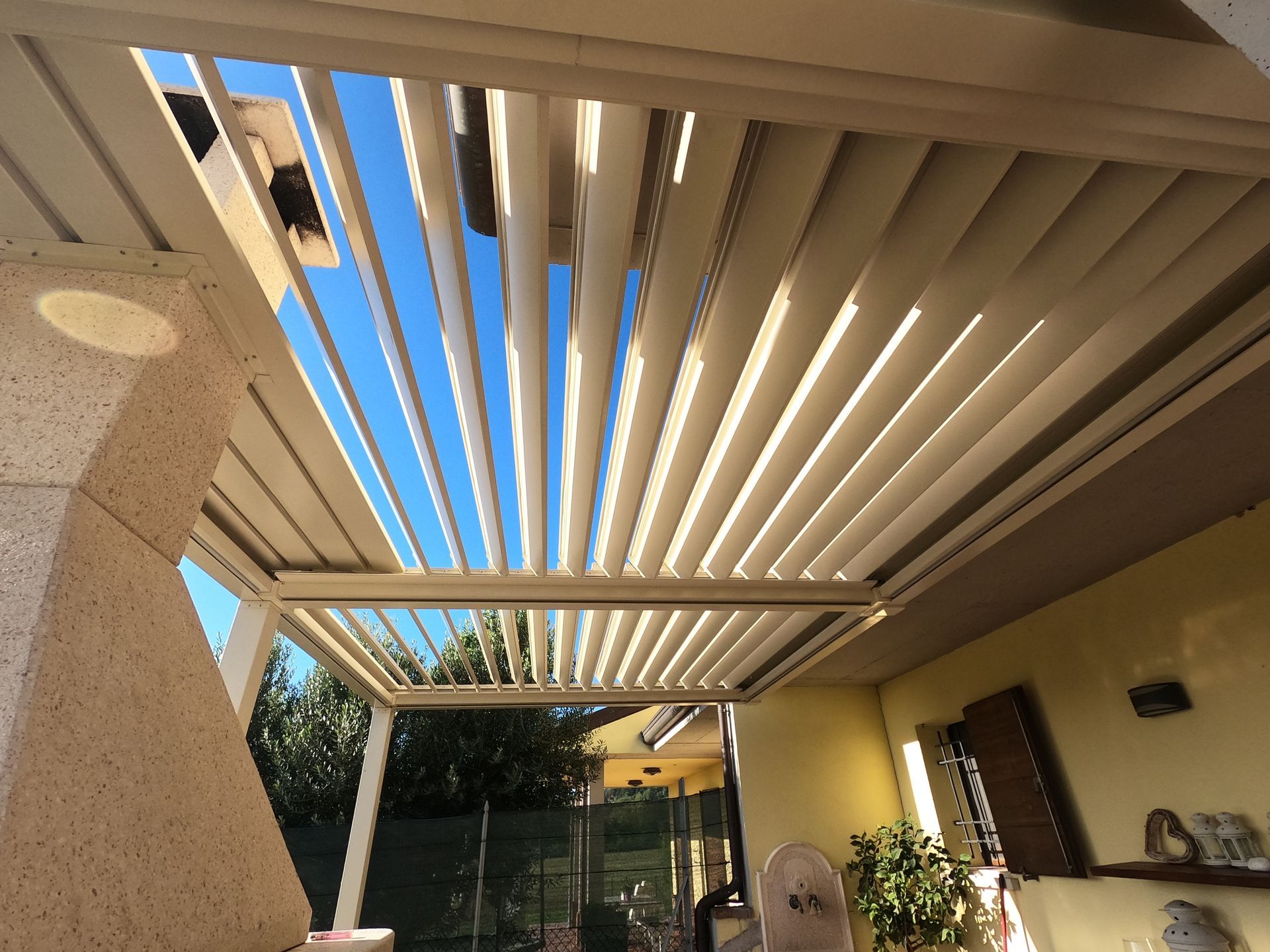 Patio coperto con pergolato a doghe beige, pareti color crema e un cielo azzurro oltre il giardino.