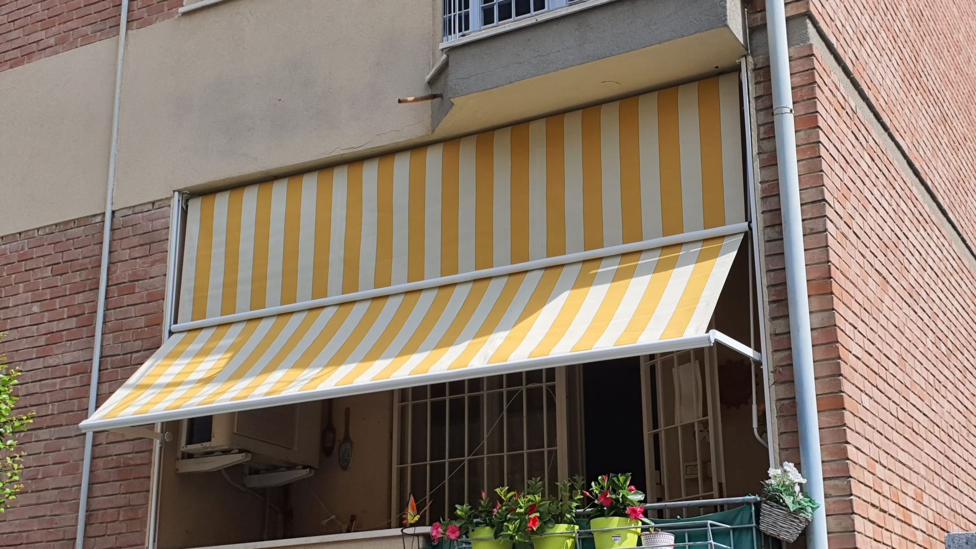 Tenda a righe gialle e bianche sopra il balcone di un appartamento in mattoni, con piante in vaso e ringhiera in metallo.