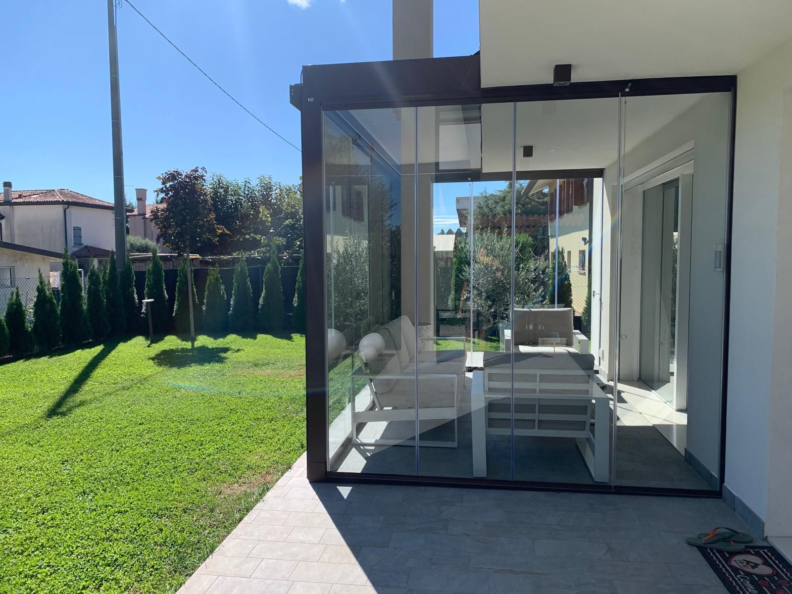 Patio vetrato con sedie bianche che si affaccia su un prato verde e soleggiato e sul giardino.