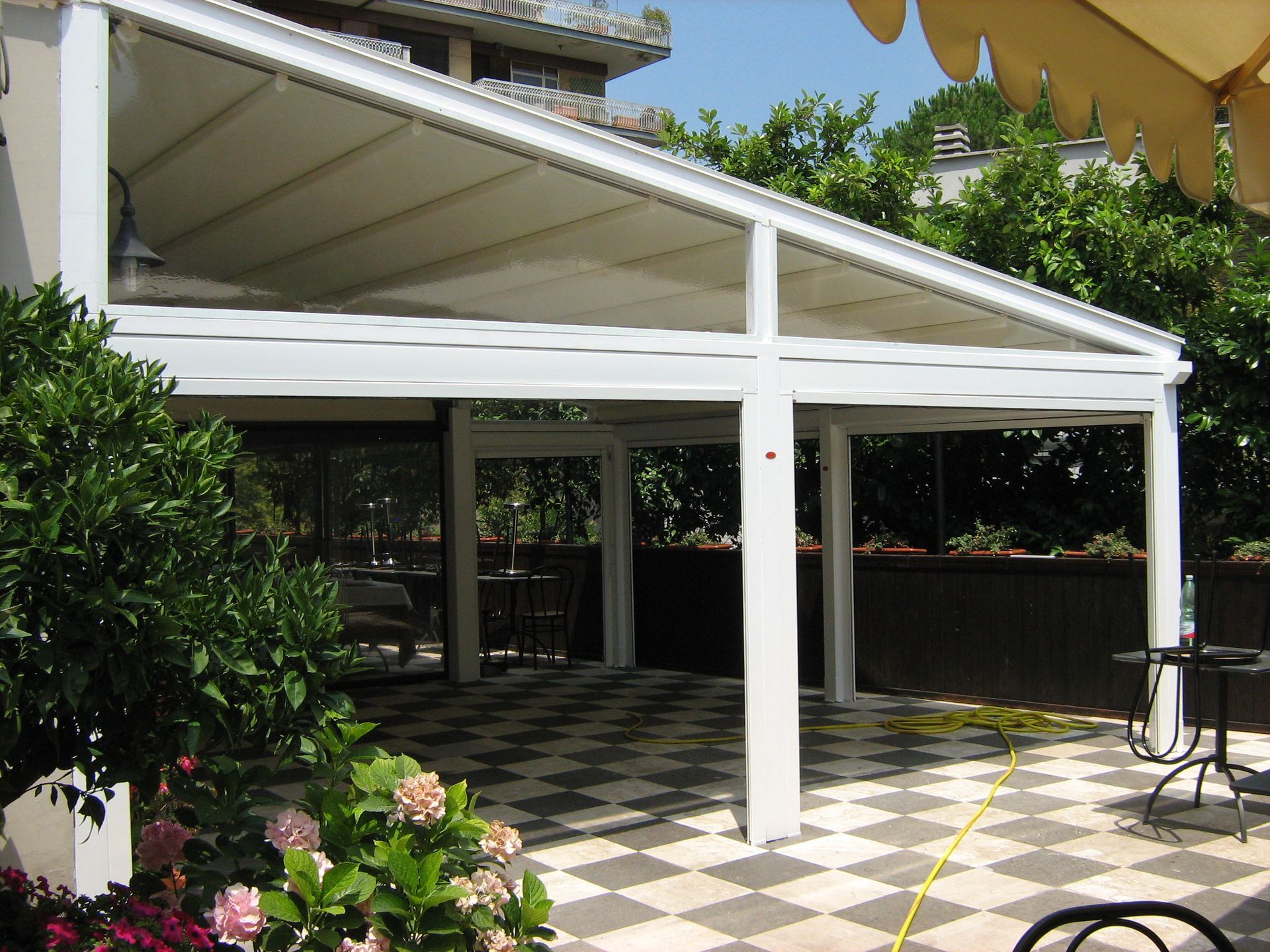 Tenda bianca da patio sopra una zona salotto esterna ombreggiata con pavimento a scacchiera e piante da giardino.