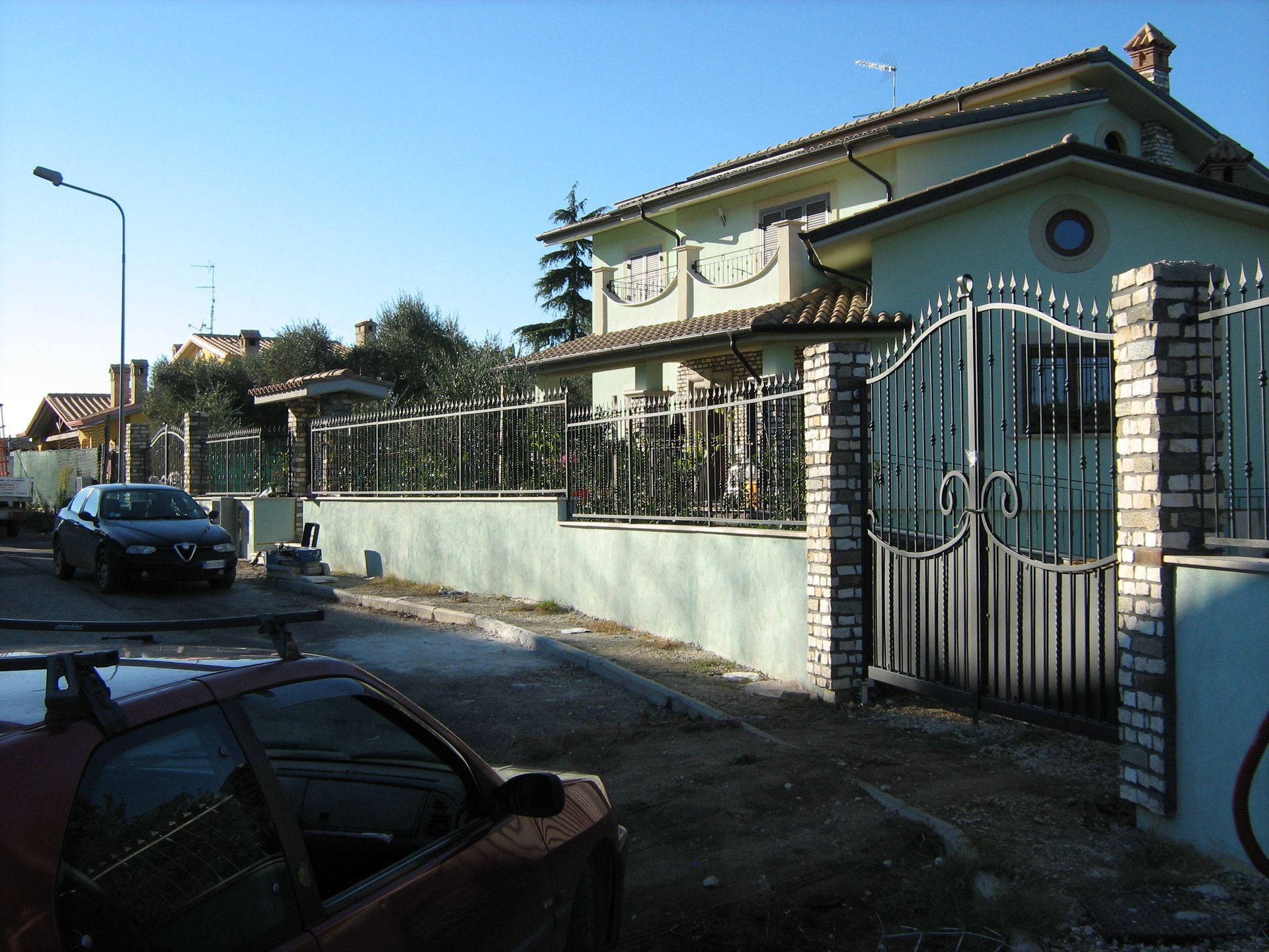 Tranquilla strada residenziale con una casa verde a due piani, cancello in metallo, auto parcheggiate e cielo soleggiato.