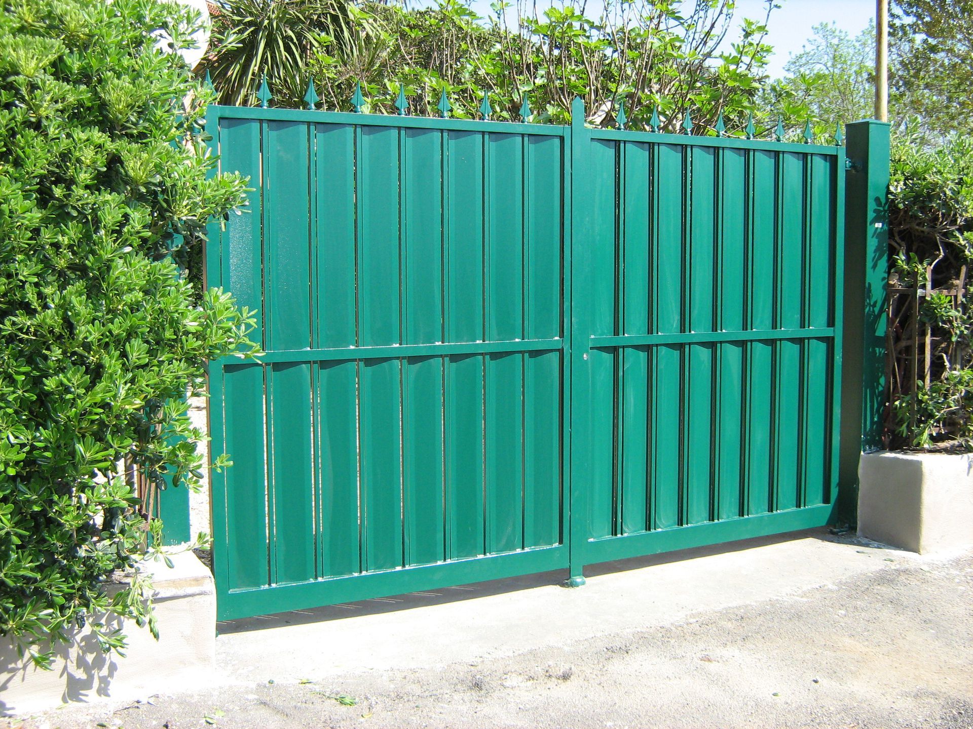 Cancello doppio in metallo verde accanto a cespugli e un muro di cemento all'esterno.