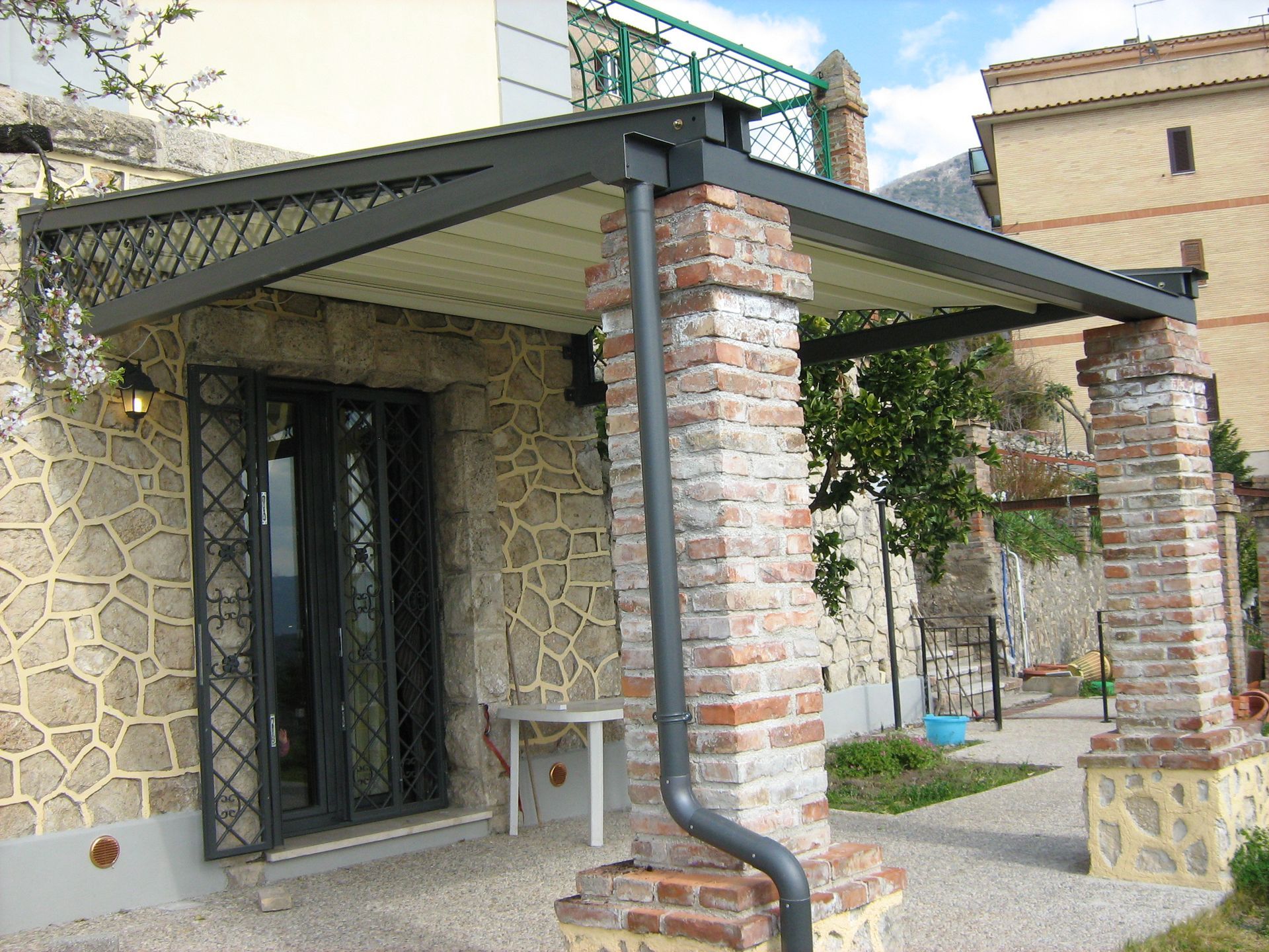 Portico in pietra con colonne in mattoni, tettoia in metallo e porta blindata nera all'ingresso di una casa