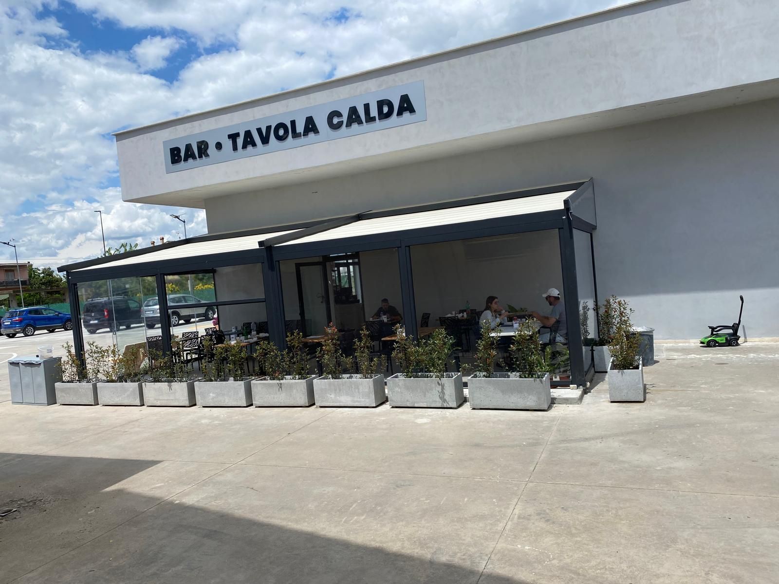 Esterno di un bar e di una taverna con piatti caldi, con patio coperto e piante in vaso sotto un cielo azzurro.