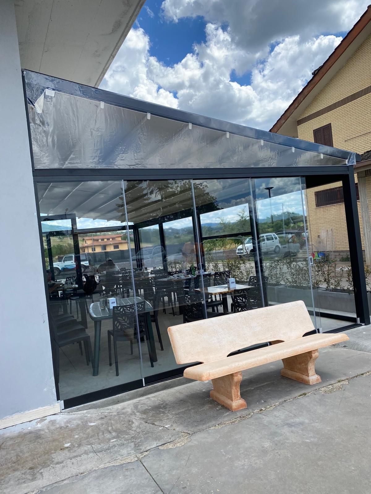 Patio vetrato con tavoli e sedie accanto a un vialetto in cemento e una panchina in legno sotto un cielo nuvoloso.