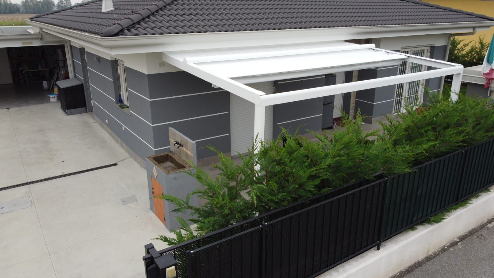 Casa moderna grigia con patio coperto bianco, vialetto d'accesso e recinzione nera, immersa nel verde.
