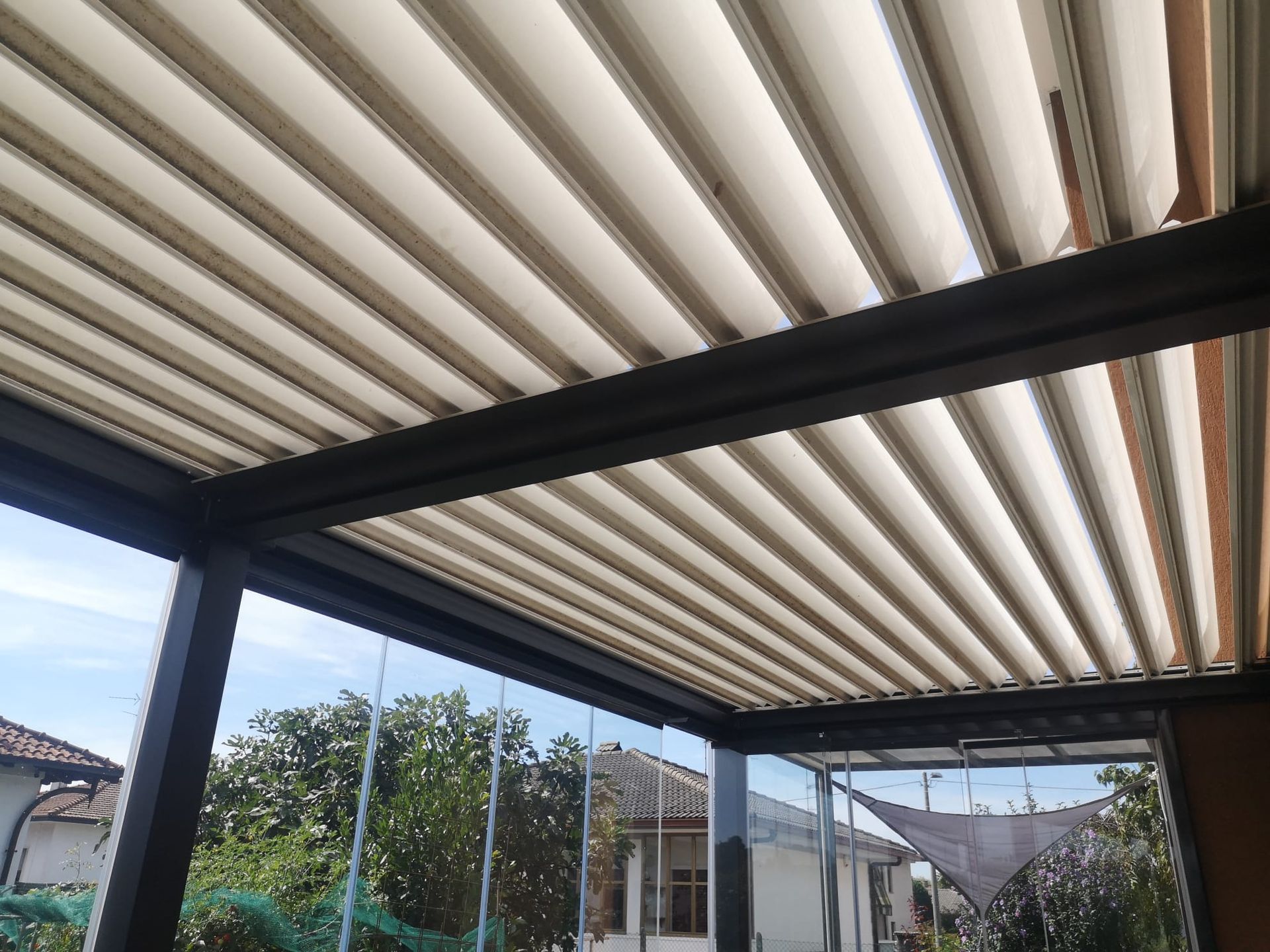 Patio coperto con tetto retrattile bianco e struttura nera, affacciato su un cortile e sulle case vicine.
