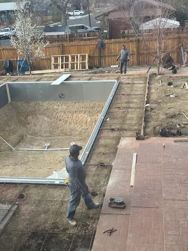 Workers install a swimming pool and wooden walkway in a backyard under construction.