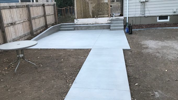 Concrete patio walkway and slab in a backyard, with a round table on dirt and a house in the background.