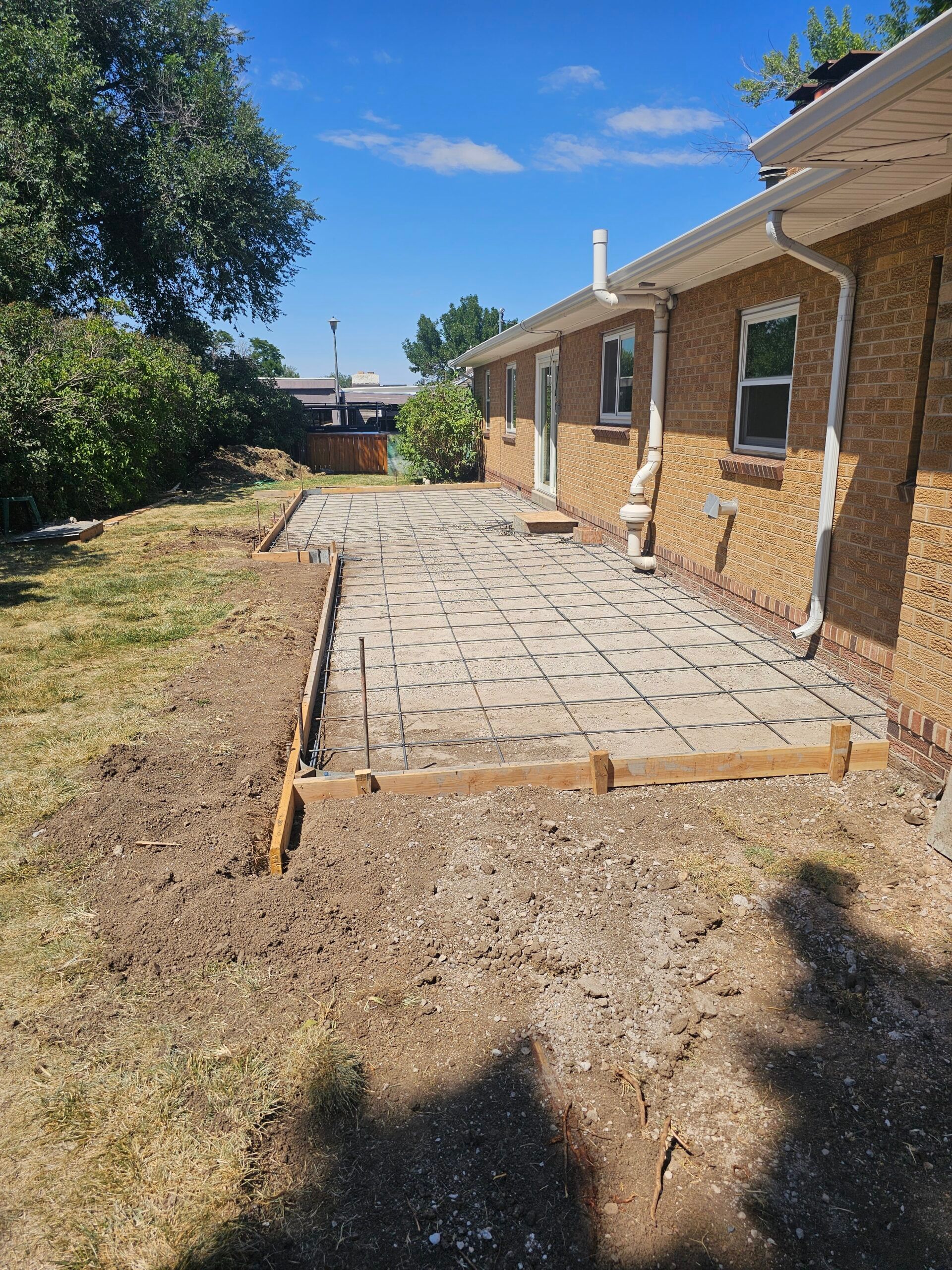 Backyard patio of gray pavers beside a brick house, with dirt and grass around it under a blue sky