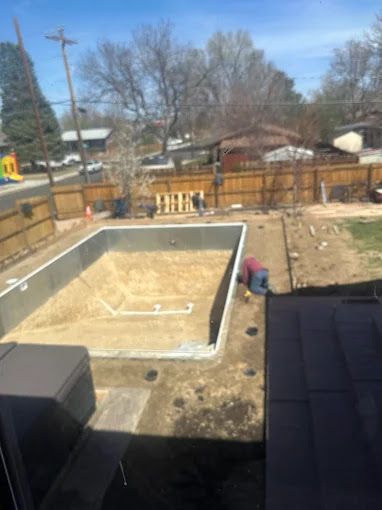 Backyard with an empty rectangular in-ground pool under construction, surrounded by dirt and a wooden fence.