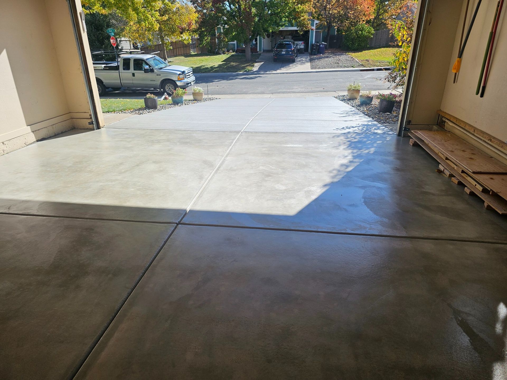 View from a garage onto a sunlit driveway and street with trees and parked vehicles.