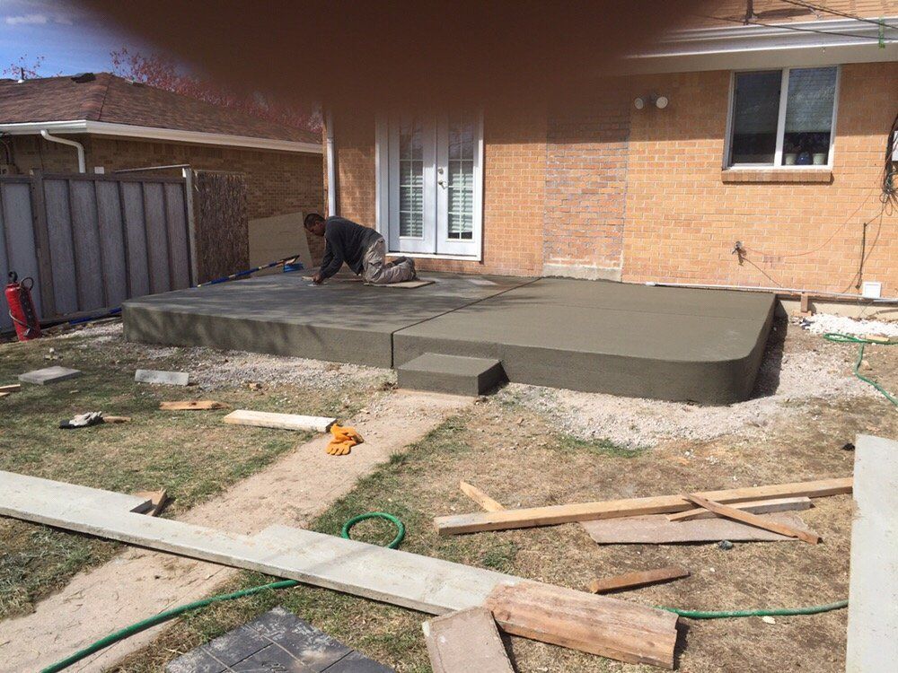 Backyard patio under construction with a worker smoothing fresh concrete beside a brick house.