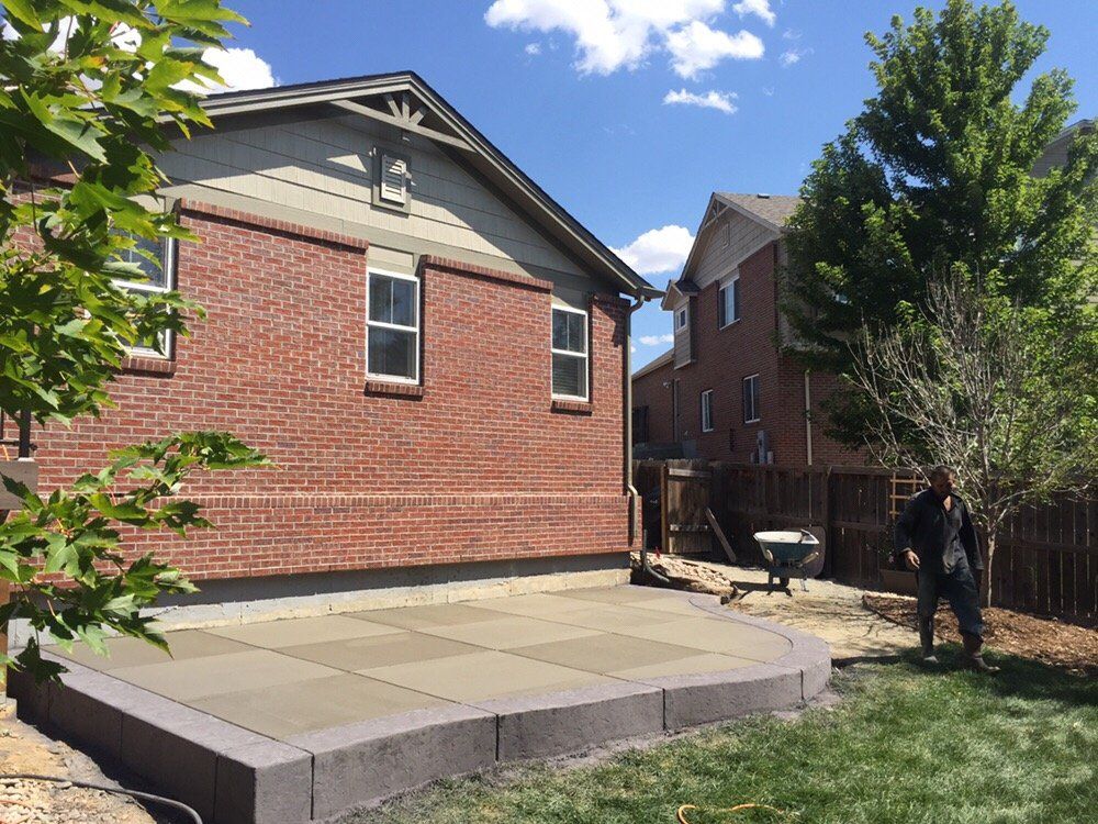 Backyard patio beside a red brick house, with a small tree and a person standing near a fence.