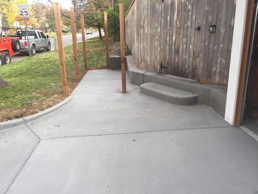 Concrete walkway beside weathered wooden building with a small step and parking area nearby