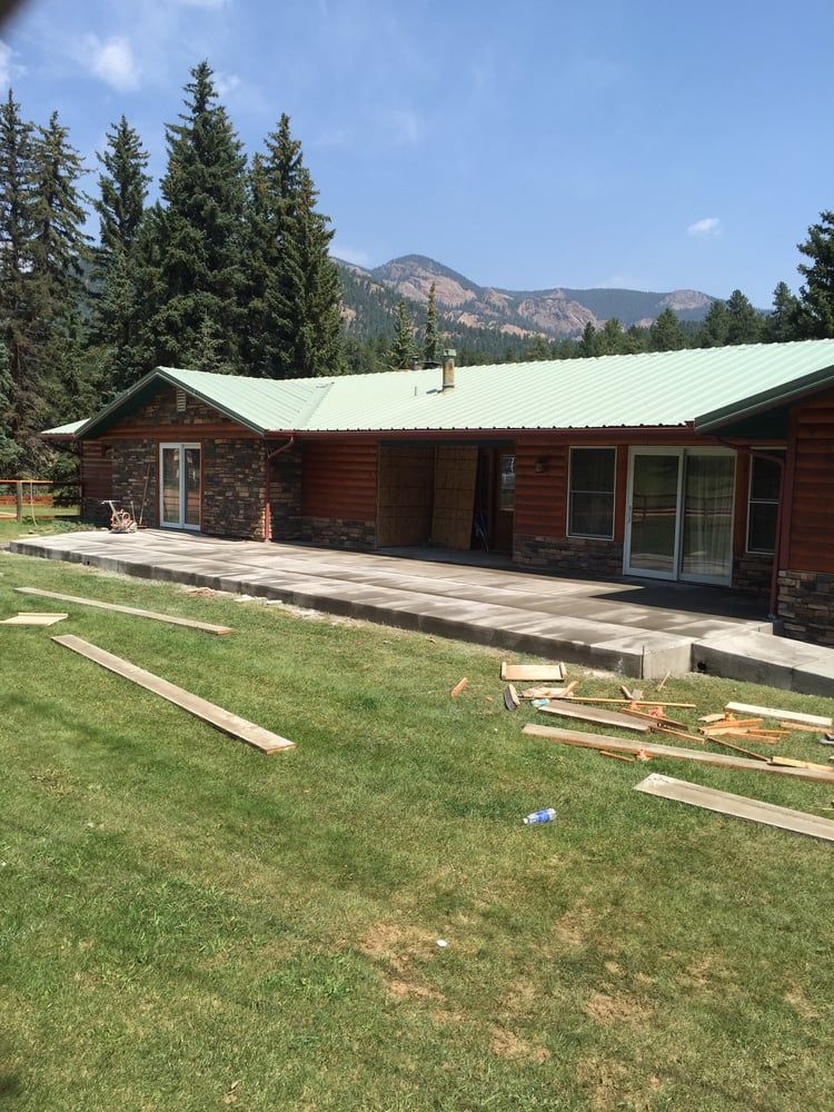 Rustic cabin with green roof beside a grassy lawn, tall pines, and mountains under a blue sky