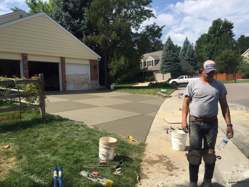 Person standing on a driveway beside buckets and tools, near a garage and freshly poured concrete.