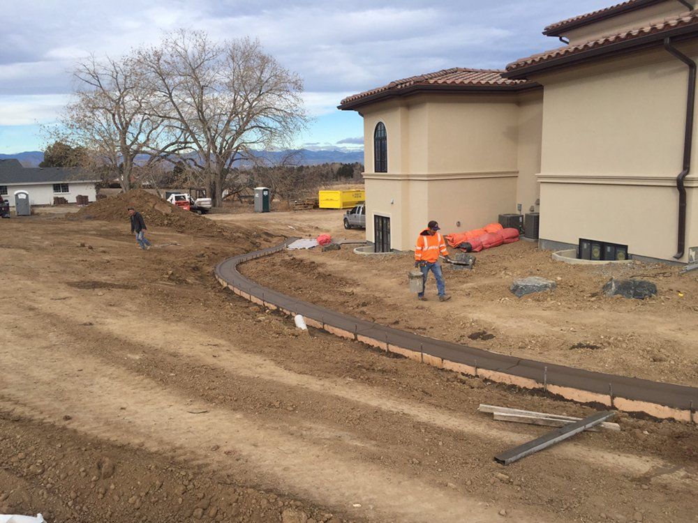 Construction workers on a dirt lot beside a beige building, with a curved curb and trees in the background.