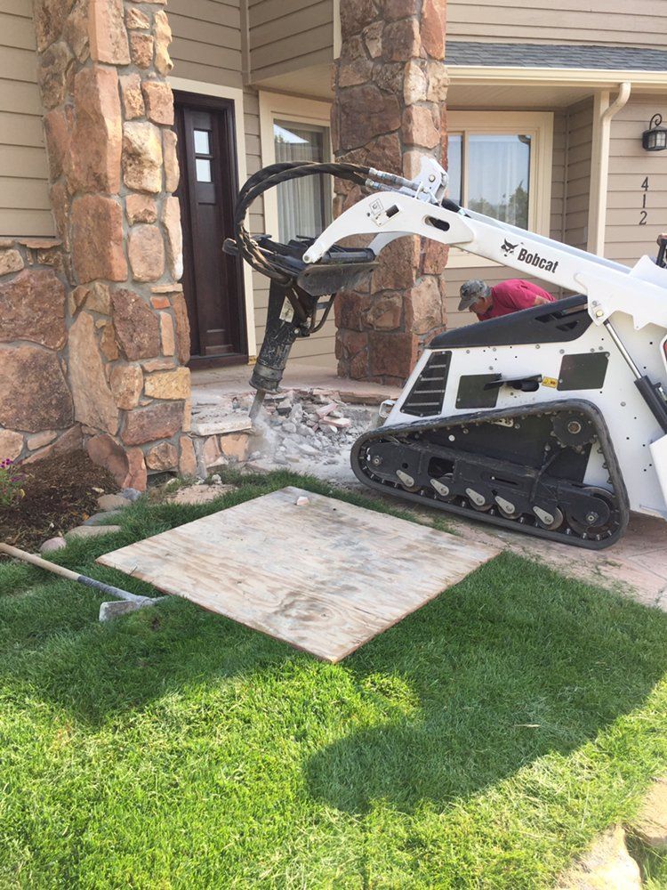White skid steer with grapple pushing a plywood sheet onto a lawn beside a house entrance